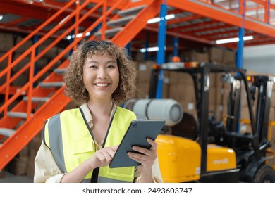 Smiling female warehouse worker in a high-visibility vest using a digital tablet, standing in a warehouse with forklifts in the background. - Powered by Shutterstock