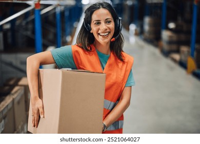 Smiling female warehouse worker stands holding a cardboard box, wearing a headset, epitomizing a blend of communication and manual handling in an industrial context. - Powered by Shutterstock