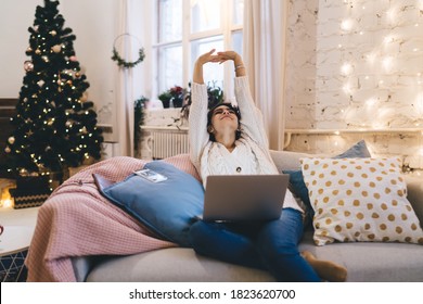 Smiling Female In Warm Sweater And Jeans Chilling On Comfortable Couch In Living Room With Laptop On Knees In Christmas Weekend