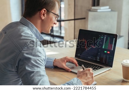 Smiling financial analyst making notes analyzing crypto currency market, sitting at his modern office table in front of laptop with candlestick chart on screen, happy with iso index growth