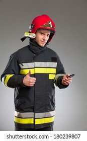 Smiling Fireman With Cell Phone Showing Thumb Up And Looking At Camera. Waist Up Studio Shot On Gray Background.