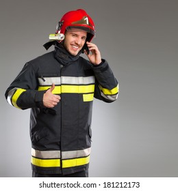 Smiling Fireman With Cell Phone Showing Thumb Up And Looking At Camera. Three Quarter Studio Shot On Gray Background.