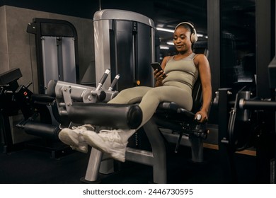 A smiling fit african american sportswoman with headphones using cellphone on leg extension machine at gym. - Powered by Shutterstock