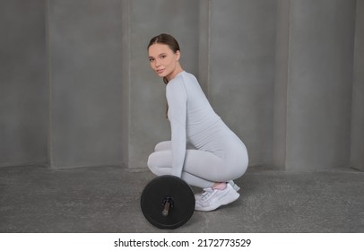 Smiling Fit Athletic Woman In Sportswear Lifting Deadlift With Barbell, Holding The Barbell, Looking At Camera And Sitting On A Grey Wall Background.

Sport And Weightlifting Deadlift Concept.