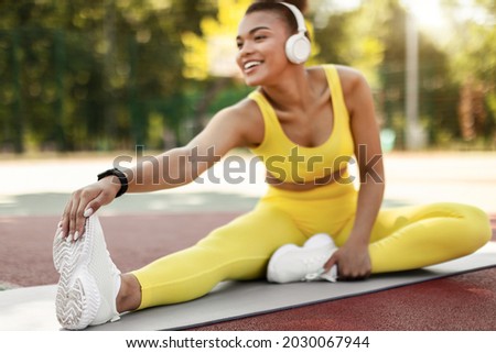 Smiling fit black woman in yellow sportswear and white sneakers stretching leg muscles sitting on yoga mat outside, touching feet, reaching toes, listening music in wireless headset, selective focus