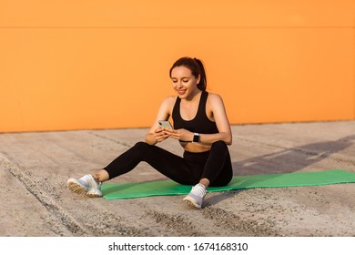 Smiling Fit Girl In Tight Sportswear, Black Pants And Top, Sitting On Yoga Mat After Workouts, Checking Body Health Indicators On Mobile Phone Application, Fitness Tracker. Sport Activity Outdoor