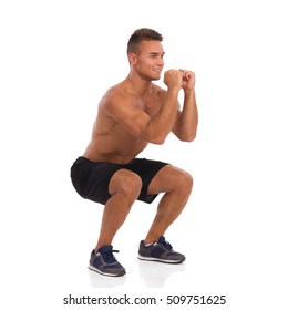 Smiling Fit Man In Black Shorts And Sneakers Showing How To Make A Squat. Front Side View. Full Length Studio Shot Isolated On White.