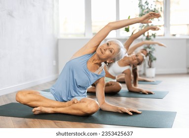 Smiling fit senior woman exercising Hatha yoga with group in modern studio, doing stretching asana Parsva Sukhasana, side bend in lotus position - Powered by Shutterstock