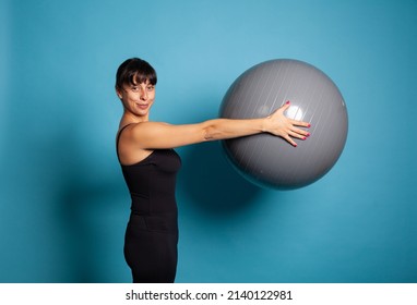 Smiling Fit Woman Holding Fitness Ball Stretching Body Muscles During Pilates Exercise In Studio With Blue Background. Athlete Person In Sportwear Working At Wellbeing. Concept Of Gymnastics