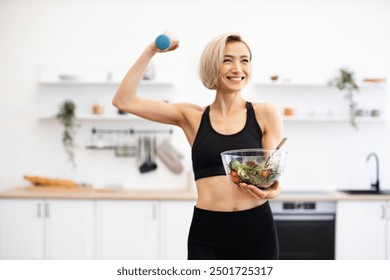 Smiling fit woman in sports bra holding salad bowl and dumbbell in modern kitchen. Embracing healthy lifestyle and fitness routine. Promoting health, nutrition, and exercise. - Powered by Shutterstock