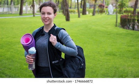 Smiling Fit Young Woman In Sportswear Holding Rolled Yoga Pilates Rubber Mat Standing In Green City Park Outdoor,looking At Camera.Fitness Instructor Sport Workout Exercise Class Training