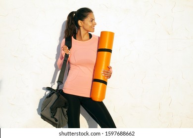 Smiling fit young woman standing on white concrete wall background, looking to the right, holding yoga mat and a gym bag, wearing black tights and pink long sleeve shirt - Powered by Shutterstock