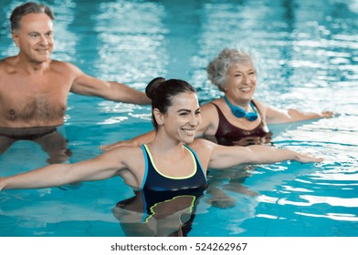 Smiling fitness class doing aqua aerobics in swimming pool. Smiling young woman with senior couple stretching arms in swimming pool. Fit mature man and old woman exercising in swimming pool.