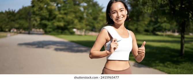 Smiling fitness girl shows thumb up, stands with towel after good workout in park, jogging and looking happy. - Powered by Shutterstock