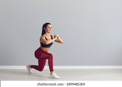 Smiling fitness trainer showing how to do fitness exercise. Happy fit young woman in sports bra and leggings doing forward lunges holding hands together in front of chest during workout at the gym
