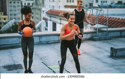 Smiling fitness women and a man doing fitness training on terrace. Man doing workout using skipping rope while his female colleagues training with agility ladder and basketball. - Powered by Shutterstock