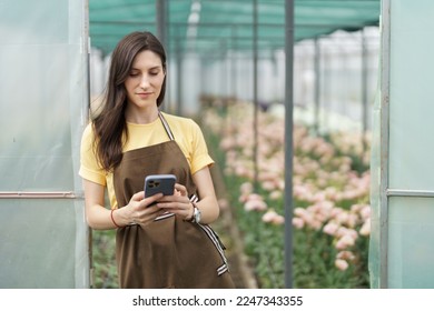 Smiling florist businesswoman using smartphone, wearing yellow t-shirt and brown apron holding phone in hands at the green house - Powered by Shutterstock