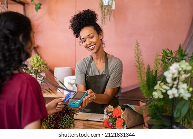 Lächelnde Floristen, die Kartenlesegerät am Schalter mit Kreditkarte bezahlen. Junge afrikanische Blumenhändler-Assistentin, die eine Zahlungsmaschine beim Kauf einer Strauchblume hält.