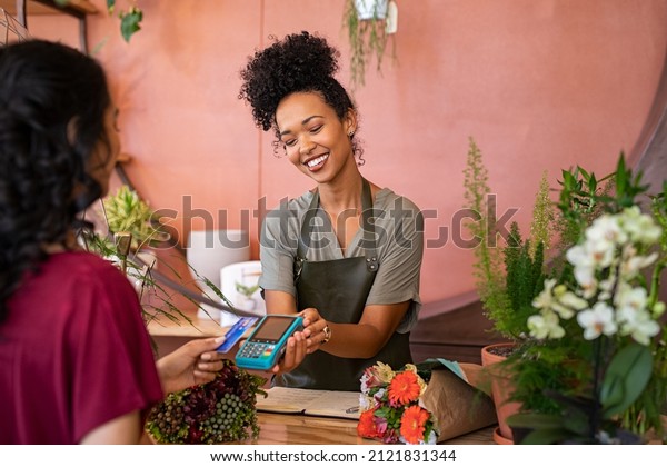 Smiling florist holding card reader machine at\
counter with customer paying with credit card. Young african\
american florist shop assistant holding payment machine while buyer\
purchase a bunch\
flower.