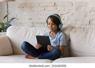 Smiling focused teenage school kid in big wireless headphones using tablet computer, relaxing on sofa at home, watching learning video, movie, attending online class, listening to music - Powered by Shutterstock
