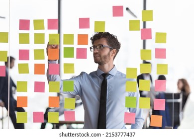 Smiling Focused Young Business Man Writing Notes On Glass Board, Working On Business Project Strategy. Startup Leader Planning Tasks For Team, Making Kanban Board, Using Scrum Management Approach