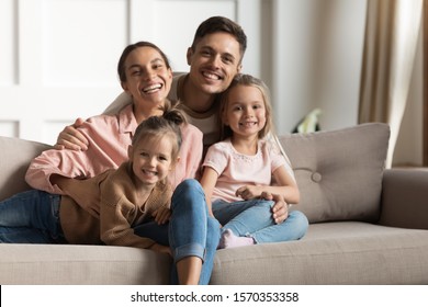 Smiling Foster Care Parents Hug Small Cute Kids Daughters Relaxing On Couch At Modern Home Together, Happy Mother Father And Children Siblings Bonding Posing For Family Portrait In Cozy Living Room