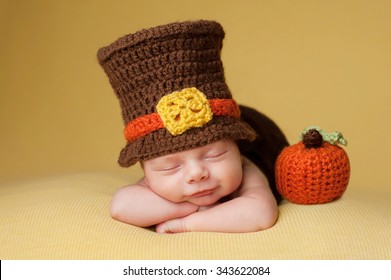 Smiling four week old newborn baby boy wearing a crocheted Pilgrim hat. He is sleeping on a gold blanket next to a crocheted pumpkin.