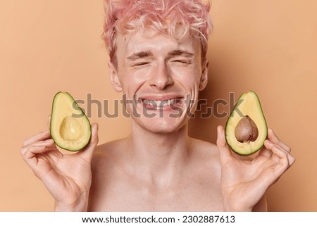 Smiling freckled man smiles happily holds halves of avocado giggles positively has healthy look has eyes closed isolated over brown background being in good mood going to undergo beauty treatments Сток-фото © 