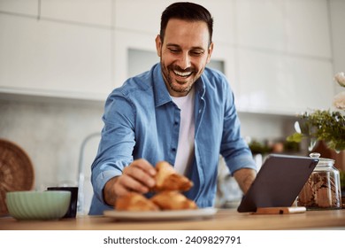 A smiling freelance man leans forward to take a croissant for a snack while standing in front of a tablet at a kitchen counter. - Powered by Shutterstock