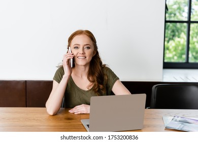 Smiling freelancer looking at camera while talking on smartphone near laptop on table - Shutterstock ID 1753200836