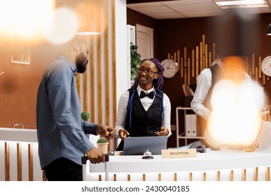 Smiling friendly African American woman receptionist talking with man guest at hotel reception. Black guy tourist standing with baggage at front desk doing check-in, receiving tourist information - Powered by Shutterstock