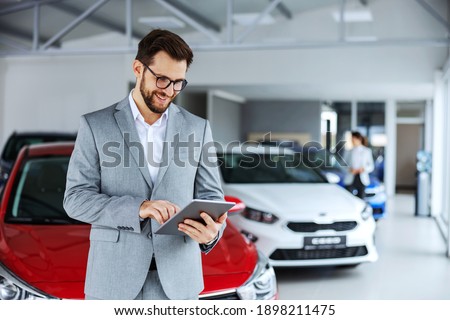 Smiling, friendly car seller standing in car salon and using tablet to check on new messages customers post on internet.