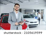 Smiling friendly car seller in suit standing in car salon and holding tablet. It