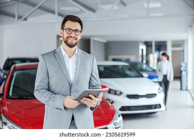 Smiling friendly car seller in suit standing in car salon and holding tablet. It's always pleasure to buy a car on a right place. - Shutterstock ID 2105619599