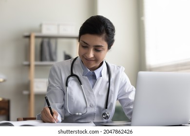 Smiling Friendly GP Doctor Giving Online Distance Video Consultation, Writing Medical Records At Laptop, Listening To Patient Complaints. Young Practitioner Watching Professional Training Webinar