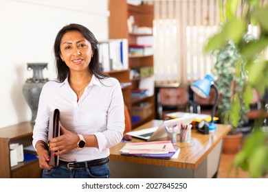 Smiling friendly Hispanic female administrative secretary standing with briefcase of documents in modern office interior.