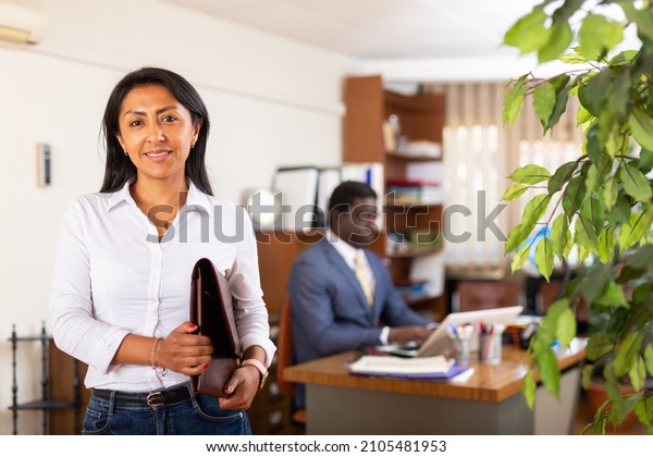 Smiling\
friendly Hispanic female administrative secretary standing with\
briefcase of documents in modern office\
interior.