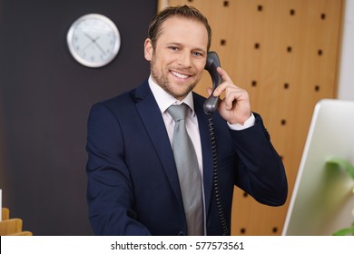 Smiling Friendly Hotel Manager Or Receptionist Taking A Call At The Front Desk In The Lobby While Standing Behind His Computer