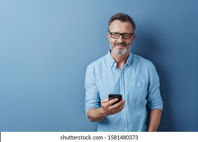 Smiling Friendly Man Wearing Glasses Standing Holding His Mobile Phone Looking At The Camera With A Warm Smile Over A Blue Studio Background With Copy Space