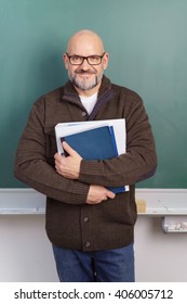 Smiling Friendly Middle Aged Male Teacher Or Senior Student Wearing Glasses Standing In Front Of A Chalkboard Clutching A File And Paperwork