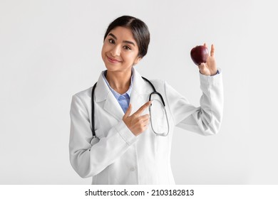 Smiling friendly millennial cute indian lady doctor in uniform shows finger on red apple isolated on white background, empty space, studio. Health care, diet, nutritional, advice from nutritionist - Shutterstock ID 2103182813