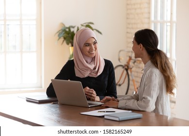 Smiling friendly mixed race colleagues communicating at workplace. Two female diverse teammates enjoying break pause time, chatting at office. Happy businesswoman discussing project ideas with client.