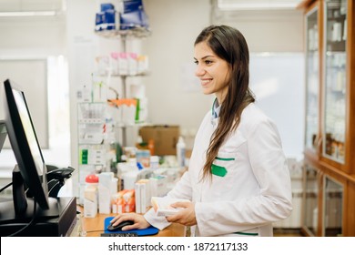 Smiling friendly pharmacist at the counter working in a pharmacy store. Pharmaceutical professional recommending an over the counter supplement. Remote patient care, working on a computer. Health care
