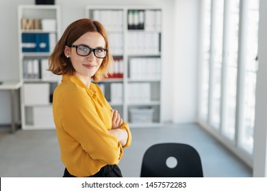 Smiling friendly self-assured young businesswoman standing sideways with folded arms in the office turning to smile at the camera