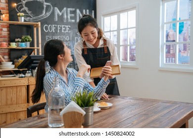 Smiling friendly waitress serving coffee drinks and croissant to friend at cafe table. cafeteria server and regular female visitors laughing together at funny joke on cellphone in modern coffeehouse. - Shutterstock ID 1450824002