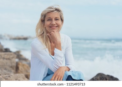 Smiling friendly woman relaxing on rocks at the beach on a misty day sitting with her chin resting on her hand looking at camera