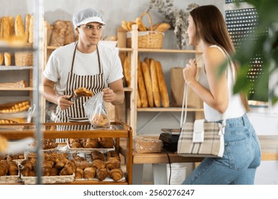 Smiling friendly young bakery worker in striped apron chatting warmly at counter and bagging fresh croissants for casually dressed female customer, surrounded by fresh bread and pastries.. - Powered by Shutterstock