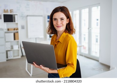 Lächeln freundliche junge Geschäftsleute, die einen Laptop auf ihrem Arm ausbalanciert halten und die Kamera ansehen, wie sie in einem hochmodernen, offenen und modernen Büro