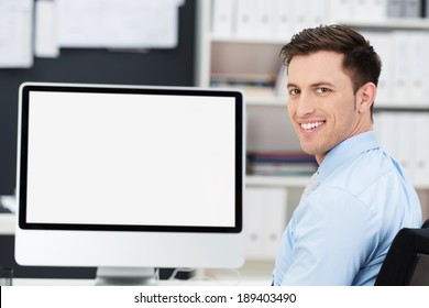 Smiling Friendly Young Businessman Sitting In Front Of A Large Blank Desktop Computer Monitor Turning To Look At The Camera, Screen Fully Visible