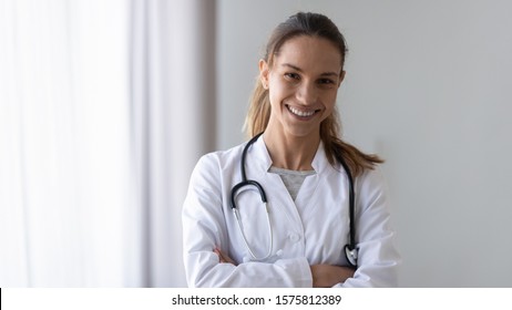 Smiling Friendly Young Female Professional Confident Doctor In White Coat With Phonendoscope On Neck Posing With Folded Hands Head Shot. Medical Insurance, Health Care, Disease Prevention Concept.